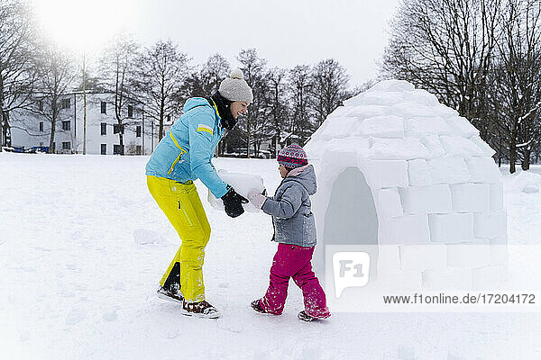Girl with help of mother carrying snow block to build igloo at park