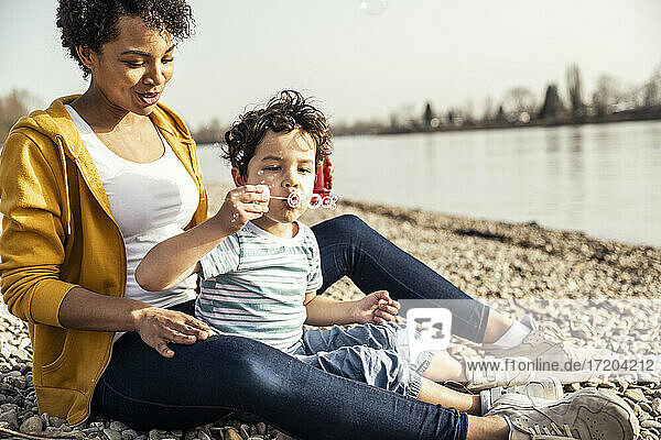 Boy blowing in bubble wand while sitting with woman on pebble