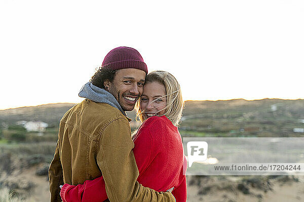 Smiling young couple looking over shoulder while standing with arm around against sky