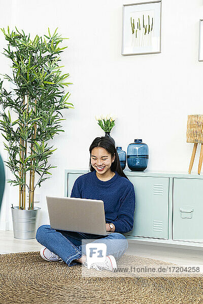 Lächelnde Frau  die einen Laptop benutzt  während sie zu Hause auf einem Juteteppich sitzt