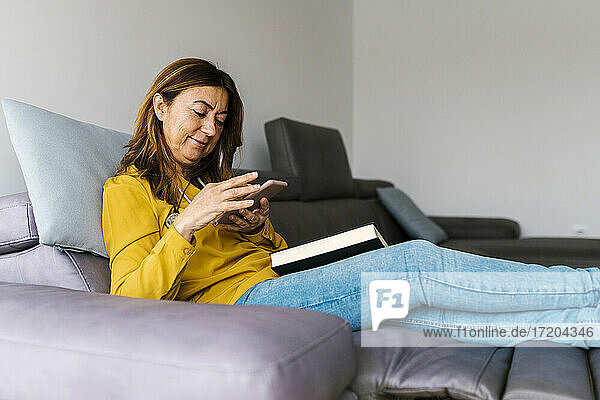 Mature woman using smart phone with book on lap while sitting on reclining chair at home