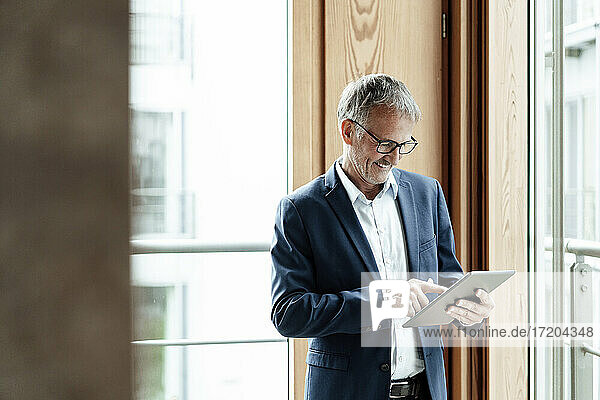 Smiling businessman using digital tablet while standing against glass window