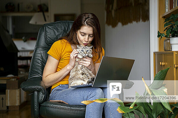 Junge Frau umarmt Katze  während sie zu Hause auf einem Stuhl mit Laptop sitzt