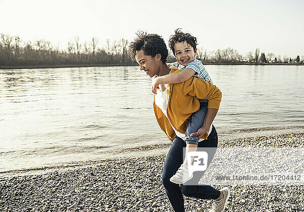 Cheerful woman piggybacking smiling boy while running by lakeshore on sunny day