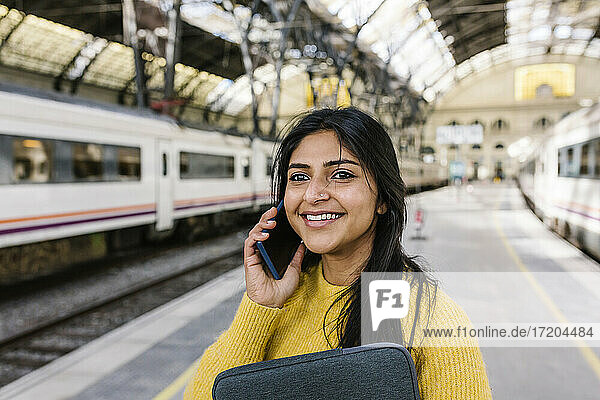 Lächelnde Frau  die wegschaut  während sie auf dem Bahnsteig mit einem Mobiltelefon spricht