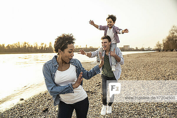 Playful father with son on shoulder running behind mother by lakeshore