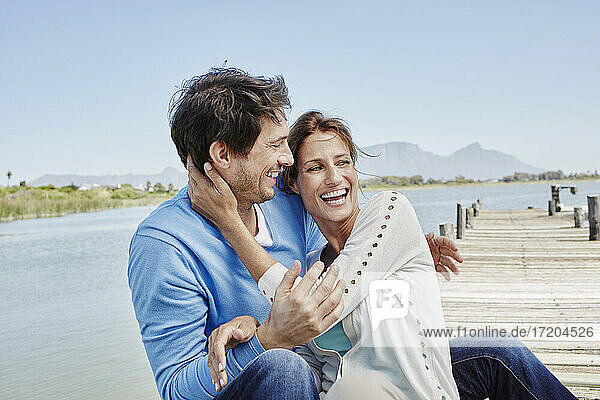 Lachendes Paar mit umschlungenen Armen auf dem Pier sitzend