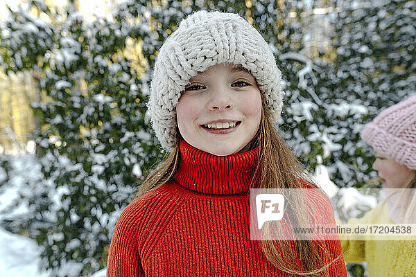 Lächelndes Mädchen mit Strickmütze  das im Schnee vor Bäumen steht