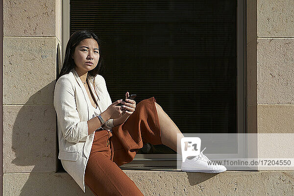 Selbstbewusste Frau mit Smartphone auf dem Fensterbrett sitzend