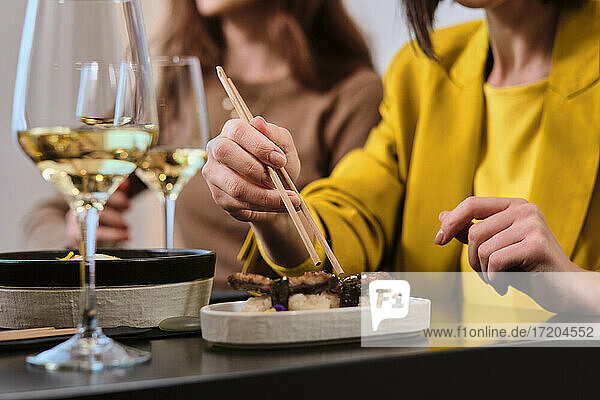 Woman eating fresh sushi with chopsticks while sitting by female friend at restaurant