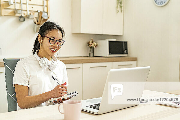 Junge Frau mit Kopfhörern  die in einen Notizblock schreibt  während sie zu Hause am Laptop arbeitet