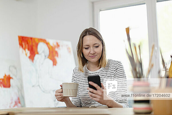 Woman having coffee while using smart phone at home studio