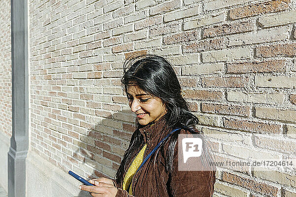 Lächelnde Frau  die ein Mobiltelefon benutzt  während sie gegen eine Ziegelmauer steht