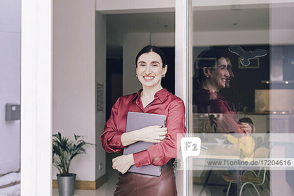 Lächelnde Geschäftsfrau  die einen Laptop hält und vor einer Glastür steht