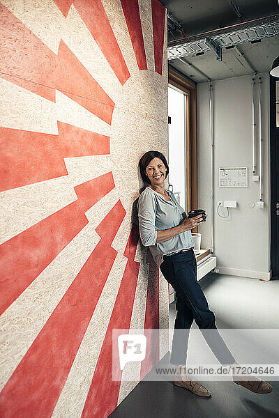 Lächelnde Geschäftsfrau mit Kaffeetasse an der Wand lehnend im Büro