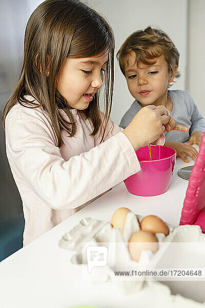 Niedlicher Junge sieht seine Schwester an  die in der Küche ein Ei in eine Schüssel schlägt
