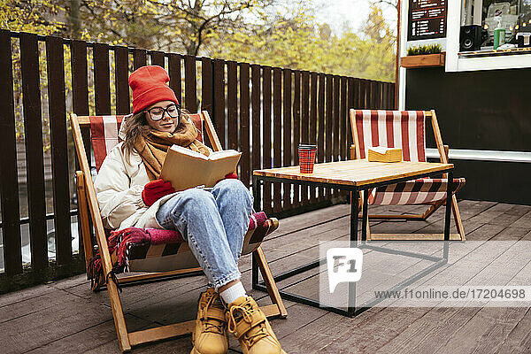 Junge Frau in warmer Kleidung liest ein Buch und sitzt auf einem Stuhl in einem Straßencafé