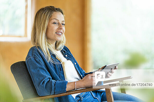 Lächelnde Frau mit digitalem Tablet  die wegschaut  während sie in einem geräumigen Zimmer auf einem Sessel sitzt