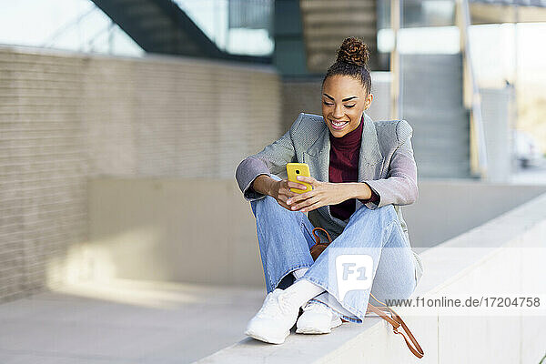 Lächelnde Geschäftsfrau  die ein Smartphone benutzt  während sie auf einer Stützmauer sitzt