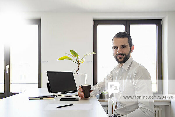 Lächelnder junger Geschäftsmann mit Kaffeetasse  der im Büro am Laptop sitzt