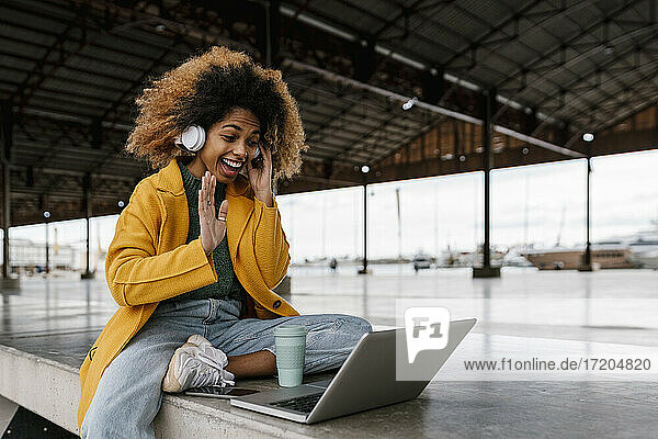 Fröhliche Afro-Frau mit In-Ear-Kopfhörern  die während eines Videogesprächs über einen Laptop im Sitzbereich mit der Hand winkt