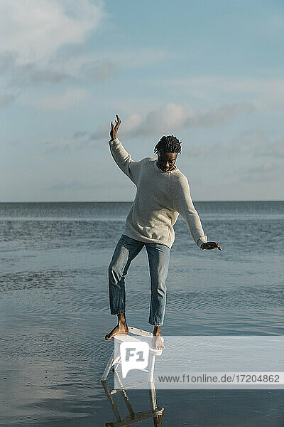 Junger Mann balanciert auf weißem Hocker am Strand gegen bewölkten Himmel