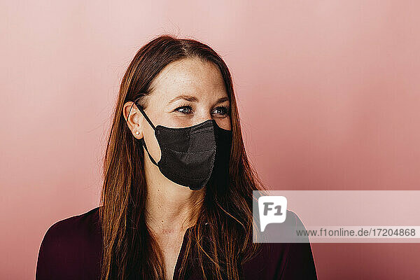 Junge Unternehmerin mit Gesichtsschutzmaske  die wegschaut  während sie vor einem farbigen Hintergrund steht