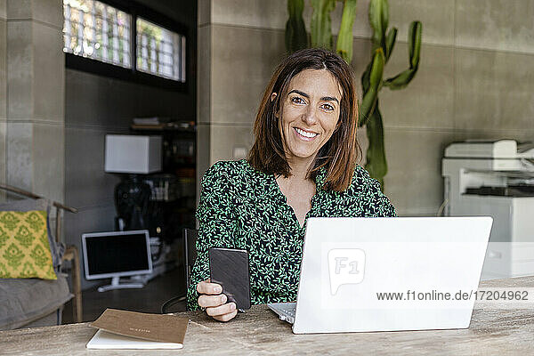 Lächelnde reife Geschäftsfrau mit Laptop sitzt am Konferenztisch im Büro