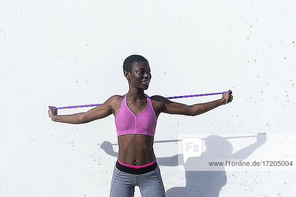Smiling female athlete stretching arms with rubber band while exercising against white wall