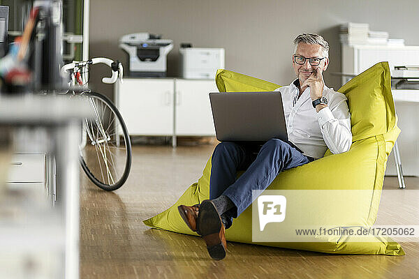 Lächelnder Geschäftsmann mit Laptop auf Sitzsack in Großraumbüro sitzend