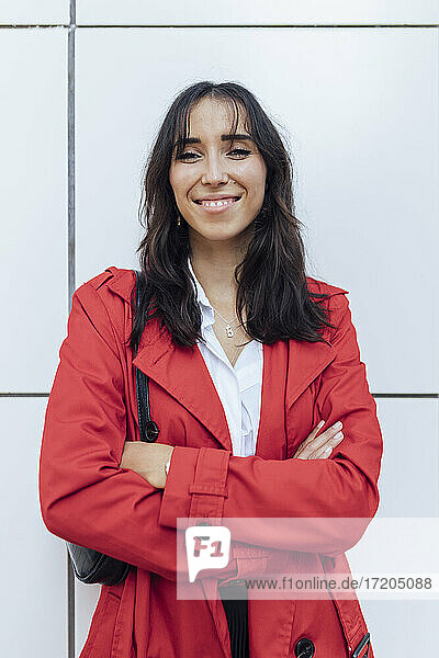 Lächelnde junge Frau in rotem Trenchcoat steht mit verschränkten Armen vor einer weißen Wand