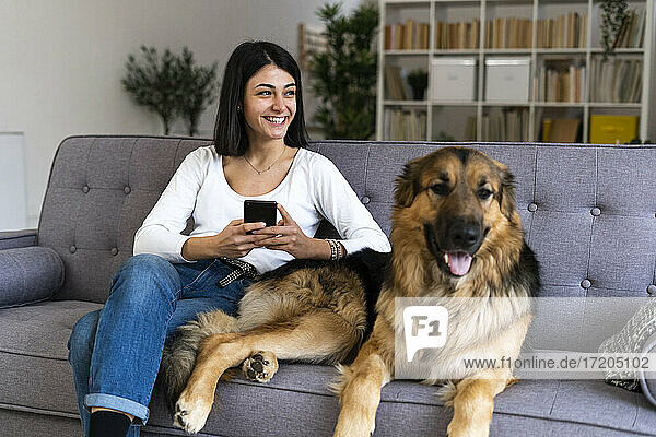 Glückliche Frau mit Smartphone  die wegschaut  während sie mit ihrem Hund zu Hause auf dem Sofa sitzt