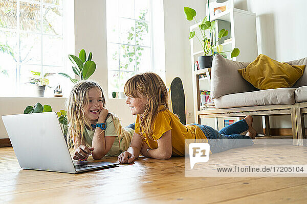 Lächelnde Mädchen  die sich gegenseitig anschauen  während sie zu Hause vor dem Laptop auf dem Boden liegen