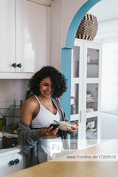 Lächelnde Frau  die eine Obstschale hält  während sie ihr Smartphone in der Küche benutzt
