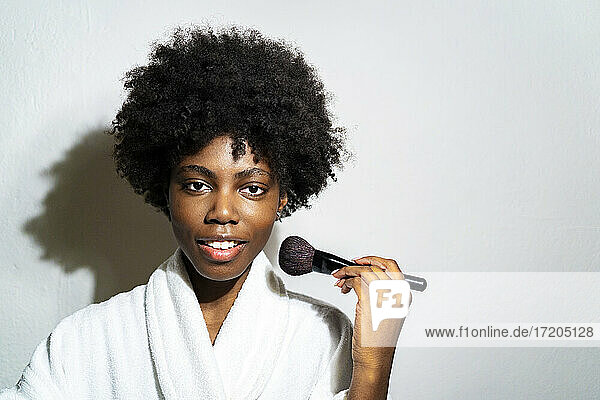 Curly woman using blushbrush while standing against white background