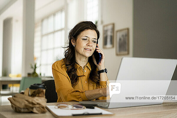 Lächelnde Geschäftsfrau  die einen Laptop benutzt  während sie am Schreibtisch im Heimbüro mit dem Handy telefoniert
