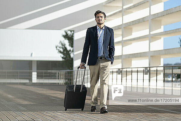 Businessman with luggage looking away while walking on footpath