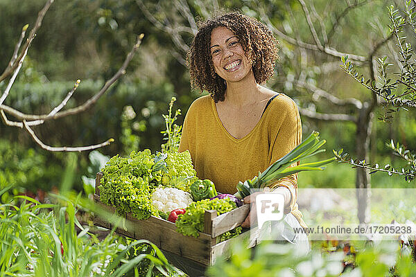 Lächelnde Frau  die eine Kiste mit frisch geerntetem Bio-Gemüse im Permakultur-Garten hält