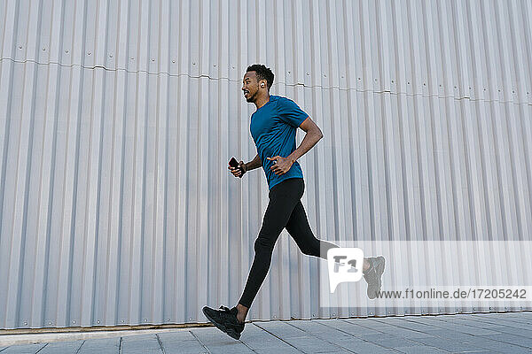 Männlicher Sportler mit Mobiltelefon  der während des Trainings auf dem Gehweg an einer Mauer läuft