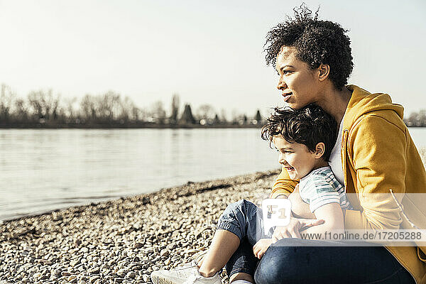Junge Mutter umarmt ihr Kind und sitzt mit ihm am Seeufer