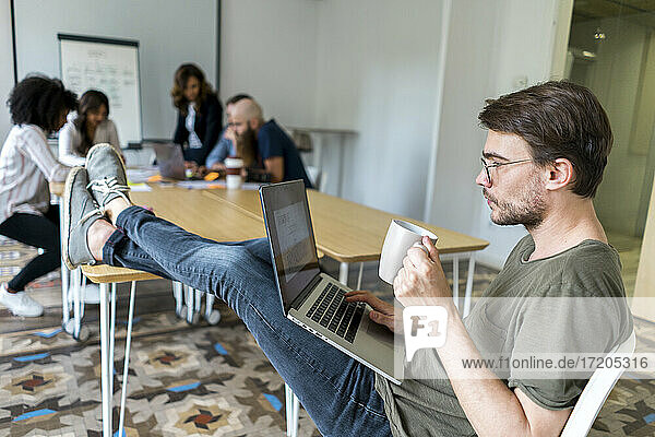 Male entrepreneur holding mug while working on laptop during meeting in office