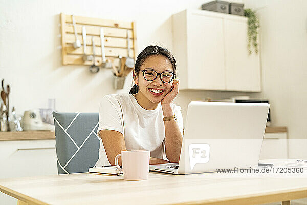 Junge Frau lächelt  während sie in der Küche am Laptop sitzt