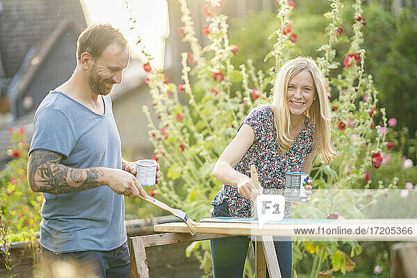 Lächelnde blondhaarige Frau  die mit einem im Garten stehenden Mann ein Holzbrett bemalt