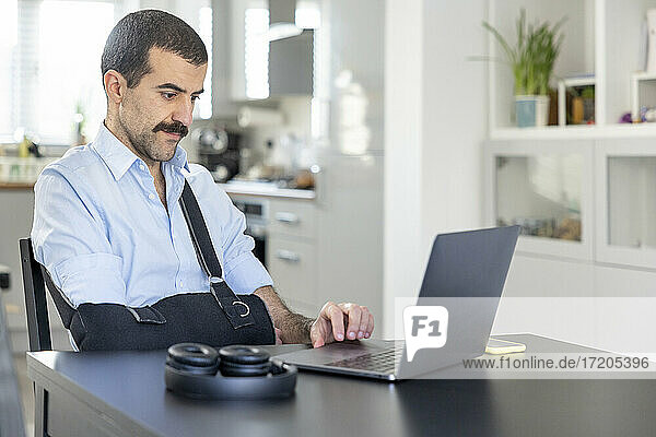 Man wearing arm sling working on laptop at home