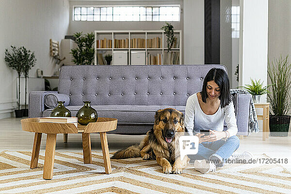 Frau  die ein Mobiltelefon benutzt  während sie mit ihrem Hund im Wohnzimmer auf dem Teppich sitzt
