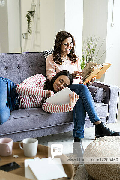 Lächelnde Tochter  die ein digitales Tablet benutzt  während sie auf dem Schoß der Mutter liegt und ein Buch auf dem Sofa zu Hause liest