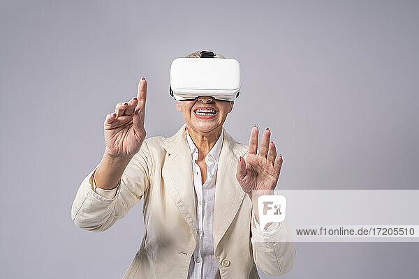 Ältere Geschäftsfrau gestikuliert  während sie ein Virtual-Reality-Headset benutzt und vor einem grauen Hintergrund steht