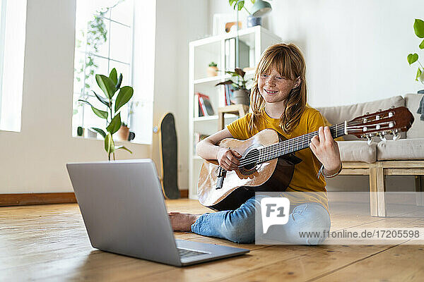 Nettes rothaariges Mädchen lernt Gitarre mit Online-Tutorial durch Laptop im Wohnzimmer zu Hause