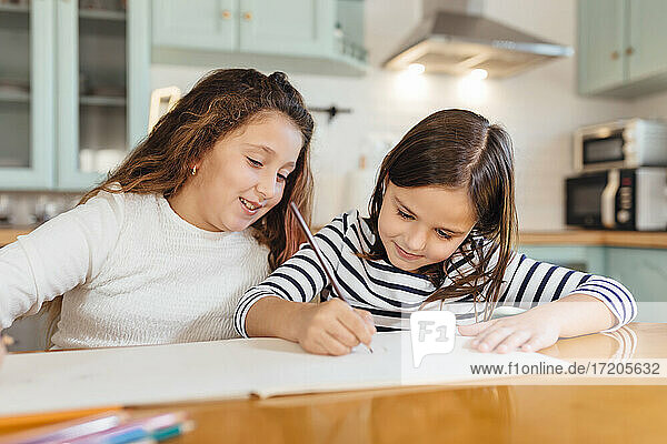 Mädchen zeichnet auf Papier  während sie mit ihrer Schwester am Esstisch in der Küche sitzt