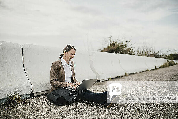Unternehmerin arbeitet am Laptop  während sie an der Leitplanke am Straßenrand sitzt